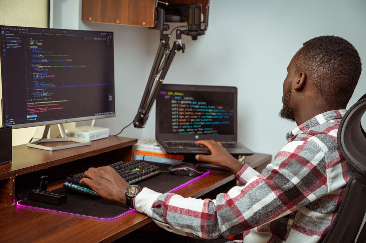 A developer in a checked shirt writes code across two monitors