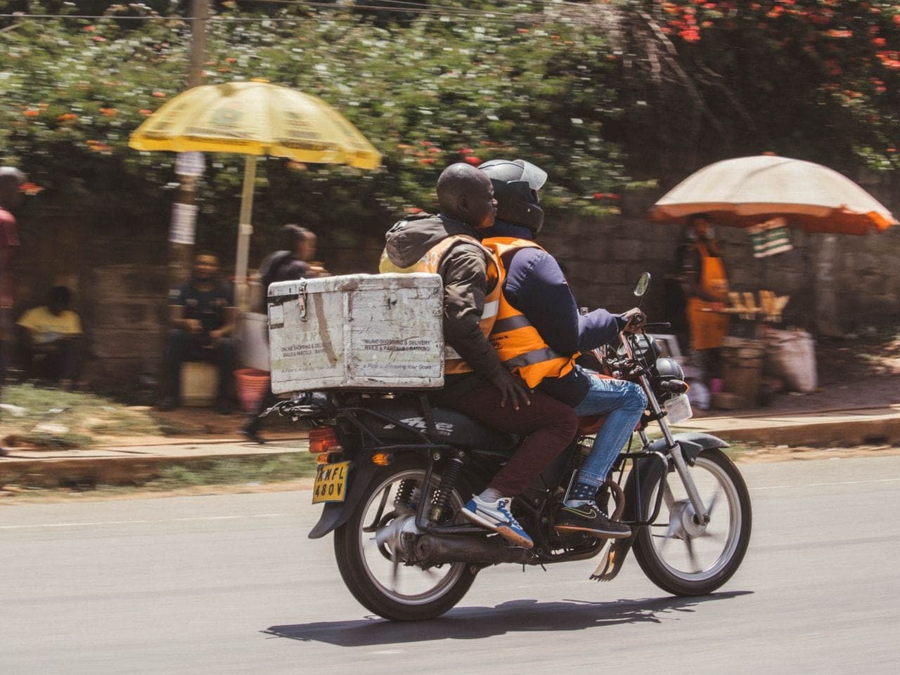 A delivery rider carries a cool box on a motorcycle along a busy street