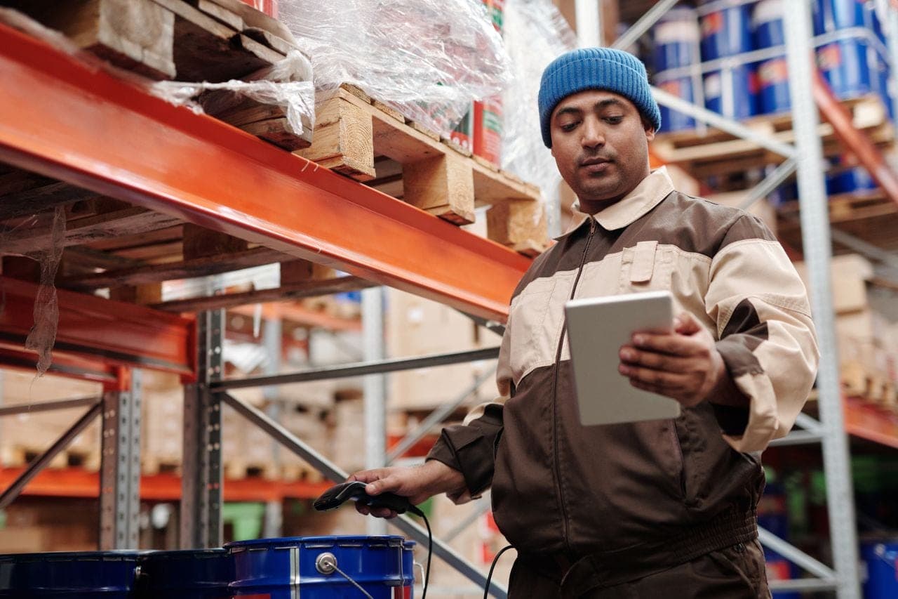 A warehouse worker in a beanie checks stock on a tablet between shelves