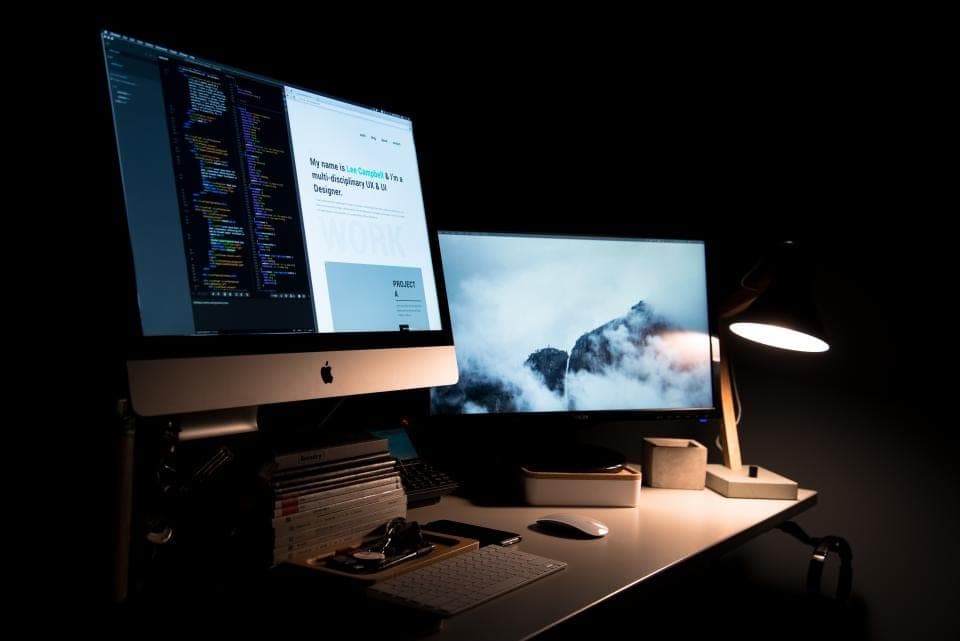 Two monitors glow on a desk in a dark room